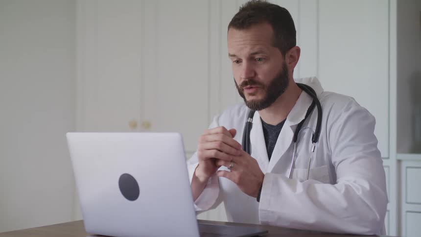 A doctor uses a laptop to speak in a video conference while sitting indoors at a table during the day