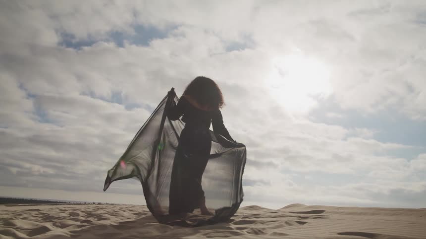 Woman wearing black walks across sand bordered by forest under cloudy sky