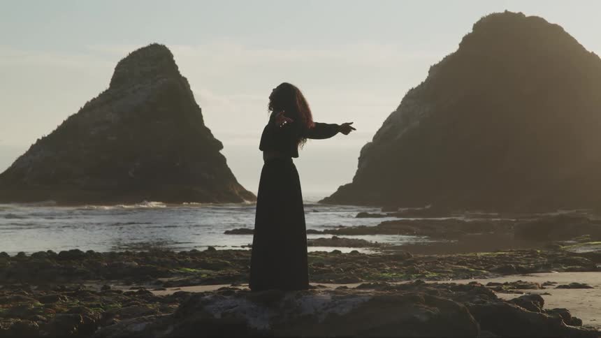 A woman in a black dress raises her arms on the beach