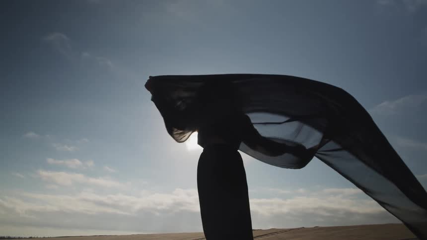Young woman wearing black dress holds a translucent cloth waving on the wind in the desert