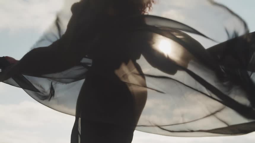 A black female in a dark dress walking in the wind with a long transparent scarf
