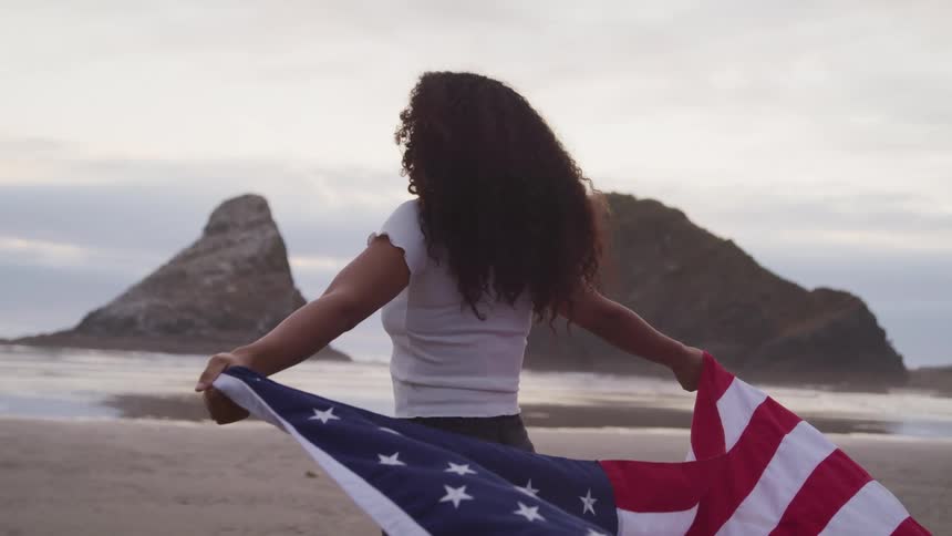 A woman stands facing the ocean with an american flag held behind her back blowing in the breeze