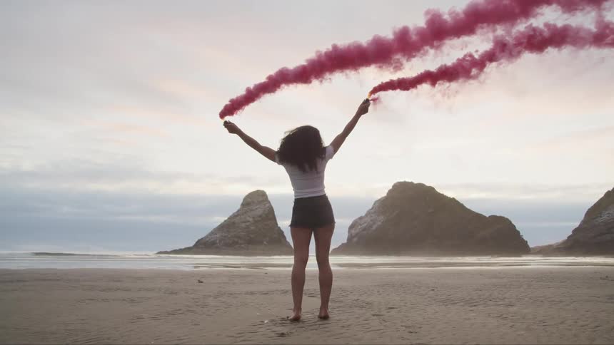 A woman with pink smoke standing on a beach with the ocean sky and clouds in the background