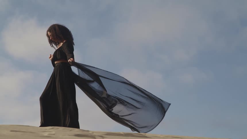Woman stands in flowing black dress at top of sand dune