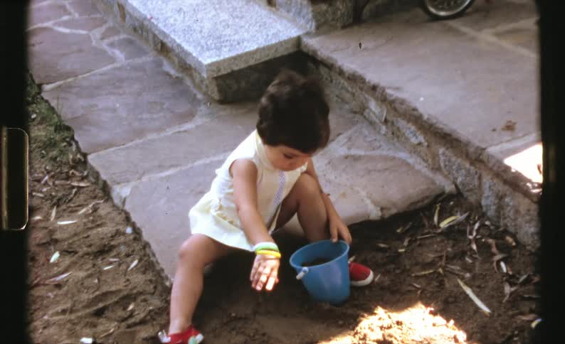 A little girl looks up then dumps out the bucket full of dirt she was playing with