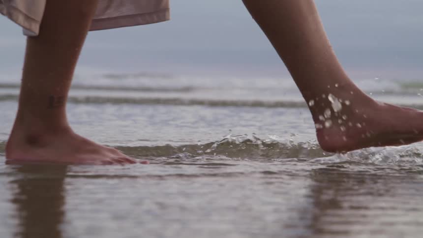 A woman with bare feet slowly walks through shallow water that has washed upon a sandy beach
