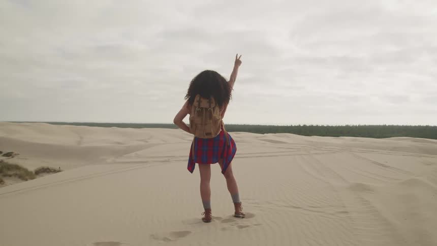 A woman standing in the sand raises her arm giving a peace sign towards the tree line