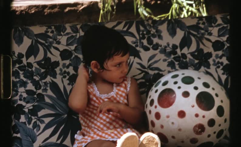 A girl sitting on a chair next to a ball with a wall and plants in the background
