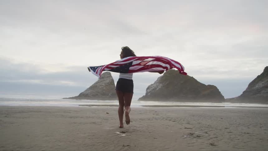 A girl is running across a beach carrying an american flag behind her