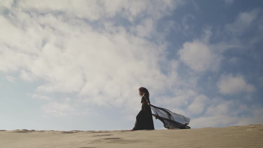 Woman walks across sand dune trailing black wrap
