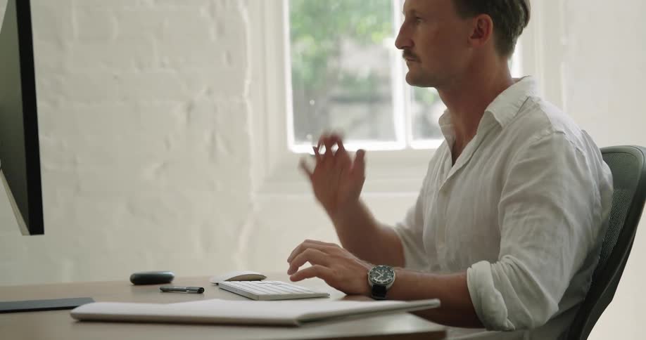 A man sits at a desk typing on a wireless keyboard near a window on a bright sunny day