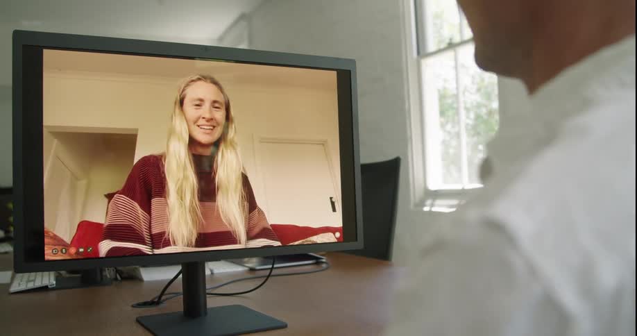 A man sitting at a desk in front of a monitor with a wall and window in the background