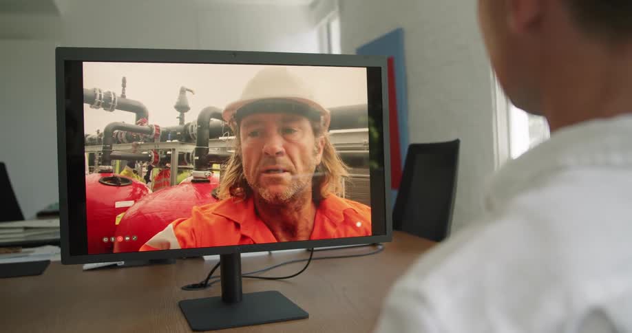 A man sitting at a conference table in an office uses video chat on a computer to speak with a man in personal protective equipment