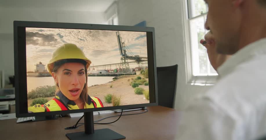 A man talks with a woman in personal protective equipment through video chat on a computer while sitting at a desk in an office during the day