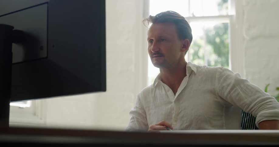 A man sitting on a chair at a desk in front of a monitor with a wall and window in the background
