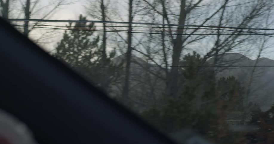 From within a car that is traveling along a road there are many trees and mountains visible in addition to telephone wires and the occasional fence