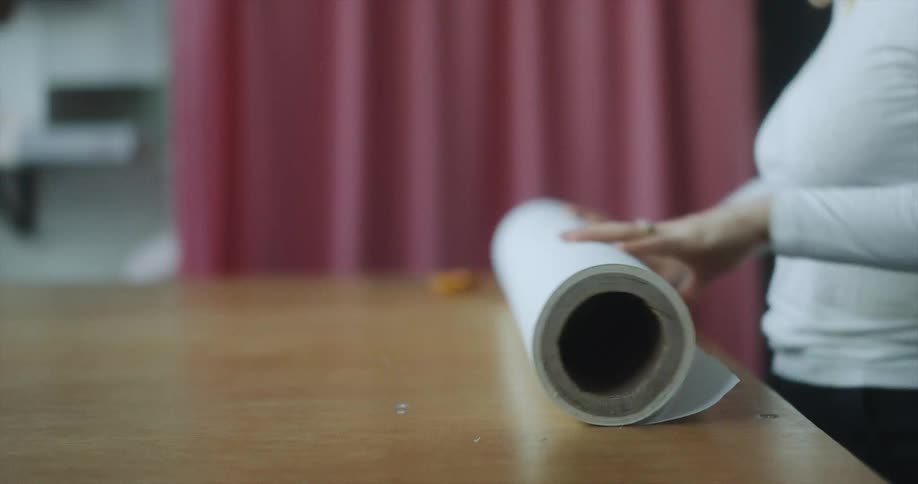 A woman stands in front of a wooden table upon which she unrolls a large roll of paper
