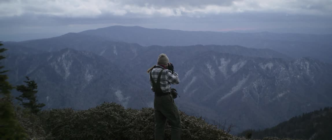 Man stands on top of a mountain and lakes a photo with a camera