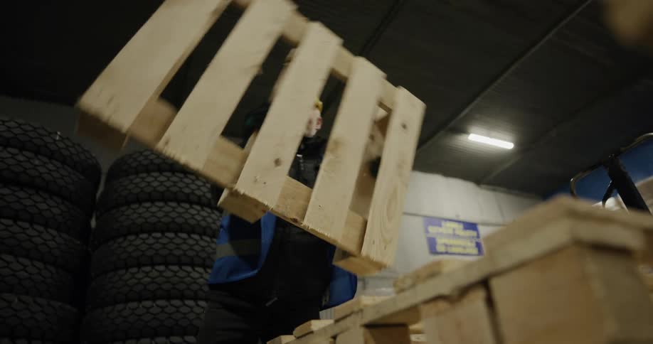 A man in personal protective equipment lifts a wooden palette and carries it out of frame