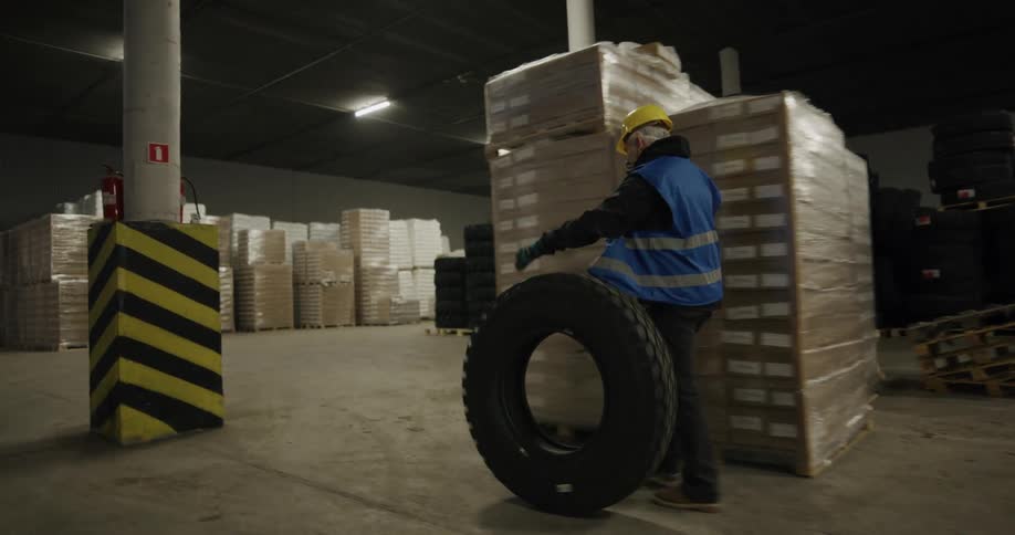 Worker wearing a yellow hardhat and blue vest rolling a tire through the warehouse