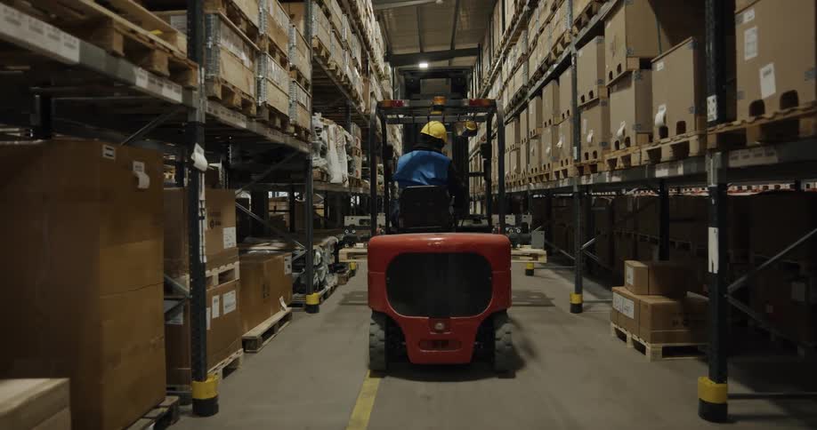 Warehouse worker drives loader along aisle past shelves full of inventory