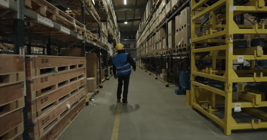 A man with a hard hat and vest walking in a warehouse with a forklift and shelves