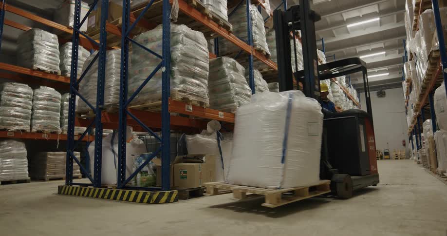 Warehouse worker drives a forklift between the aisles filled with palletes of products inside a warehouse