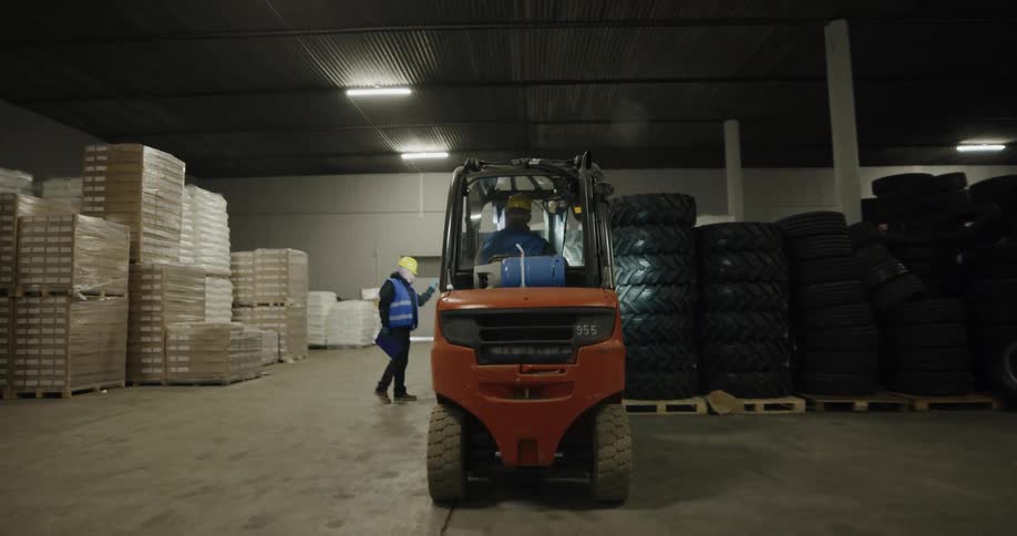 A man with a vest and hardhat operating a forklift moving tires in a warehouse