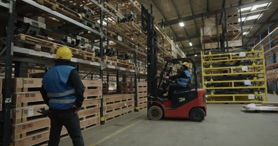 Warehouse worker uses a forklift to pick up pallets of products from a tall aisle
