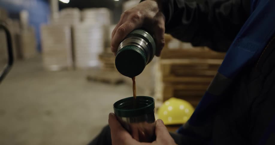 A person in a warehouse pouring coffee from a thermos into a cup