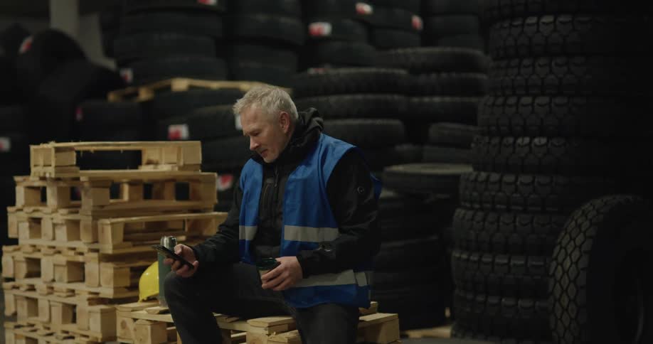 Man sits on wooden pallets taking coffee break and scrolling on mobile phone