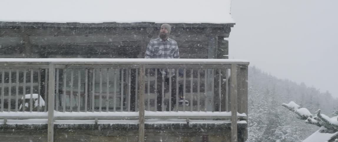 A man stands on a cabin balcony in a snowstorm