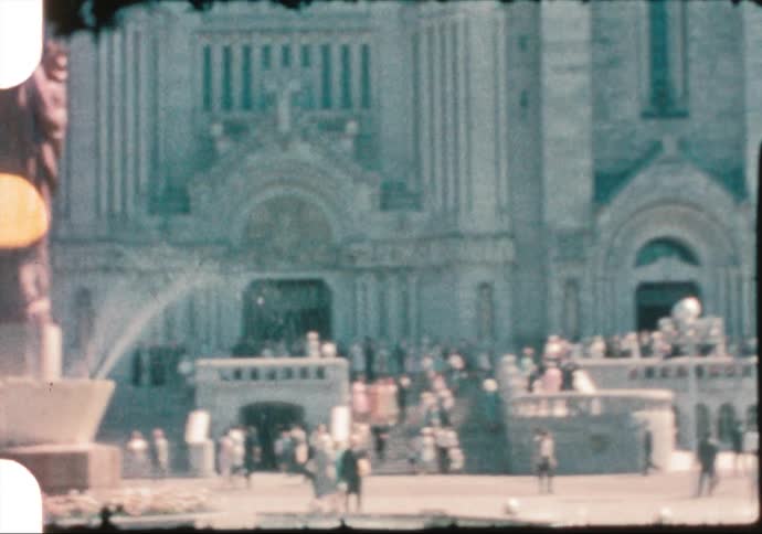 People walking in front of a church with a fountain and a statue in the foreground and the sky and clouds in the background