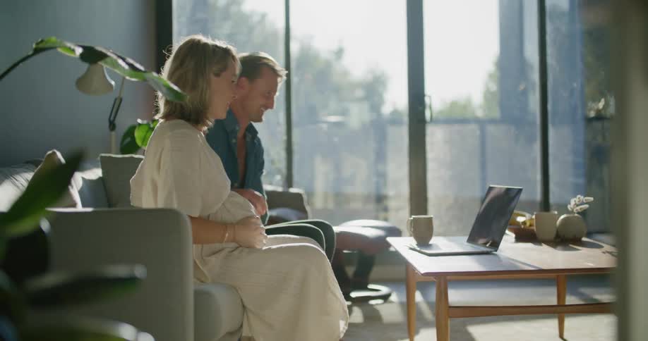 A man and woman at home hold hands and rub her pregnant belly while talking with someone on a laptop
