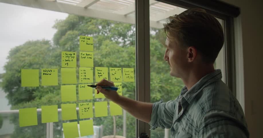 A man in a room using a marker to write on post it notes on a window