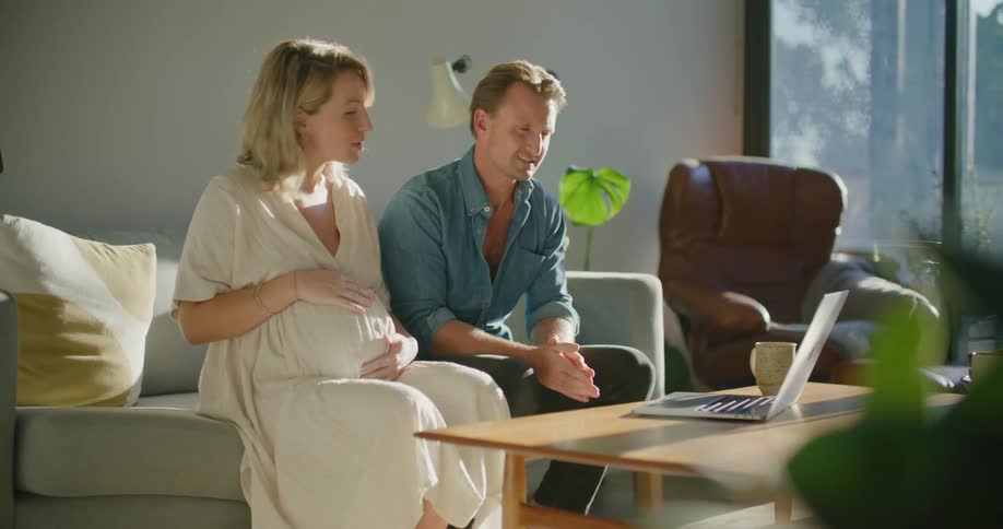 Expectant parents sit in living room holding consultation via video on a laptop