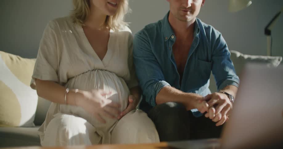 A woman rubs her pregnant stomach and smiles as she and her husband sit on the couch and talk to someone via video call on a laptop