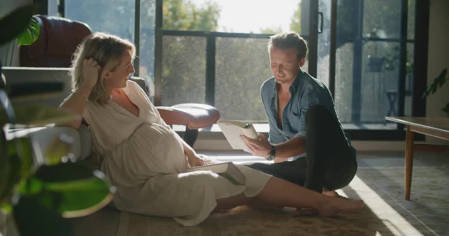 A man and woman sit on the floor at home and he reads a book to her as she rubs her pregnant belly