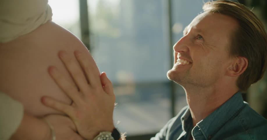Man kisses the belly of his pregnant wife in the living room at sunset