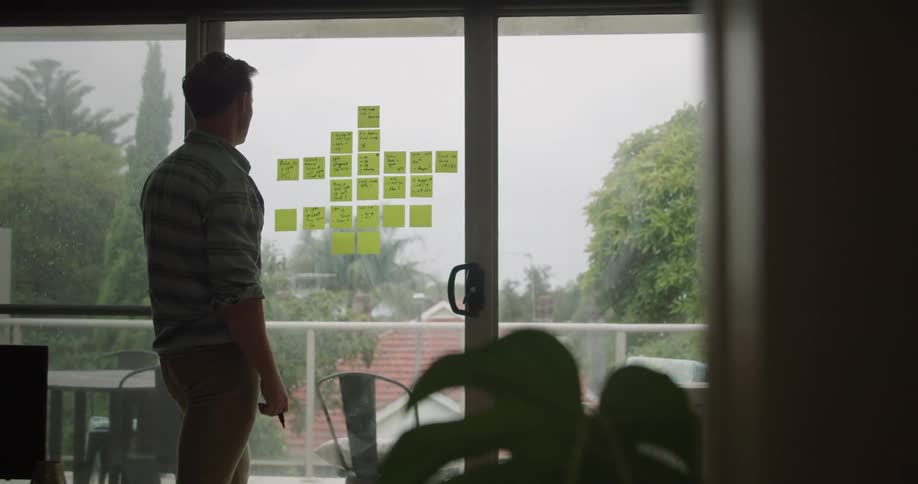 A man uses a marker to write on sticky notes attached to a large window in a dark room on a cloudy day
