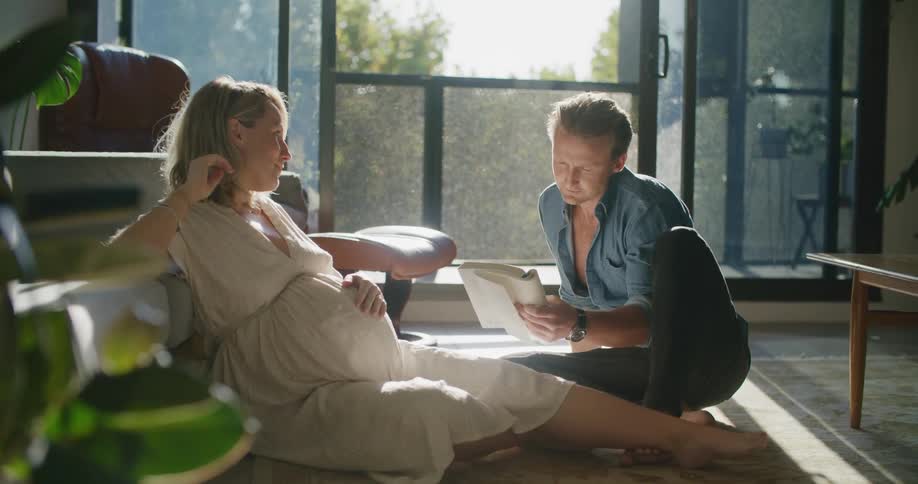 A man shows a book to a pregnant woman as they chat together while sitting on the floor of the living room of a house during the day