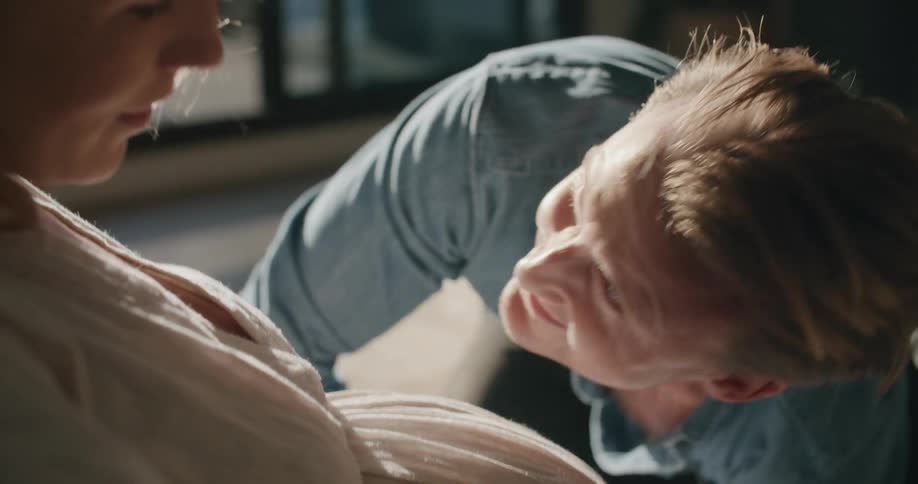 A man leans back up and smiles after listening to his wifes pregnant stomach