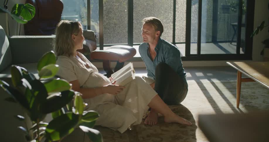 A pregnant woman reads a book while talking with a man as they sit together on the floor of the living room of a house during the day