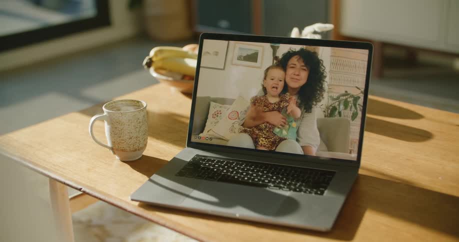 A man waves at his laptops screen while talking to a woman holding her toddler daughter on a video call