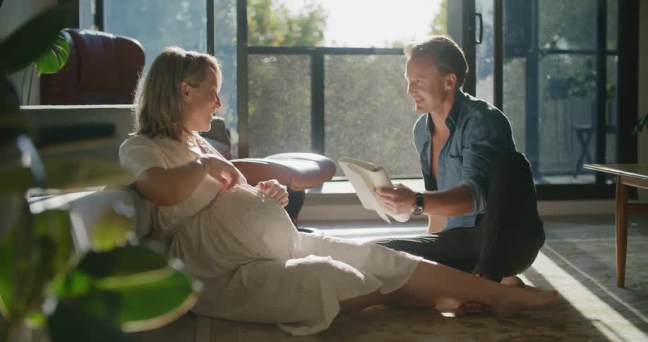 A man and woman as they sit on the floor of their condo and touch her pregnant stomach while reading a book