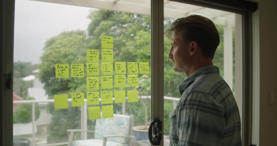 Man makes notes on post it notes stuck on sliding glass door of house as traffic passes by in distance