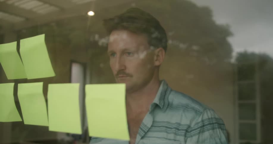 A man puts post it notes up on the inside of an office window and the glass reflects the trees and buildings outside