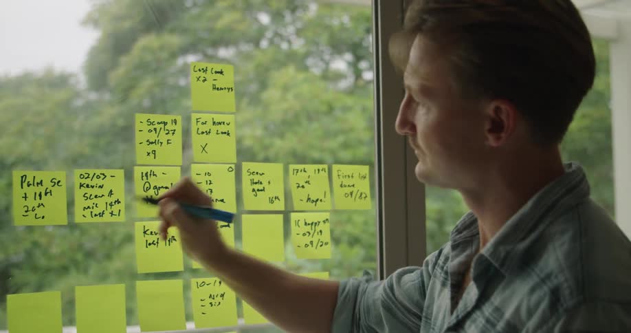 A man writes on sticky notes stuck on a large window indoors during the day