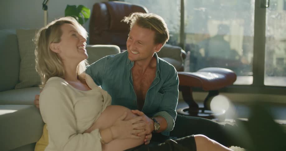 Man kisses the belly of his pregnant wife sitting in the livingroom at home