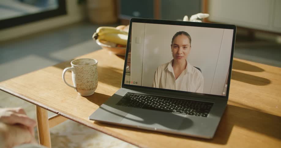 A woman on a laptop screen waves hello and smiles as she talks